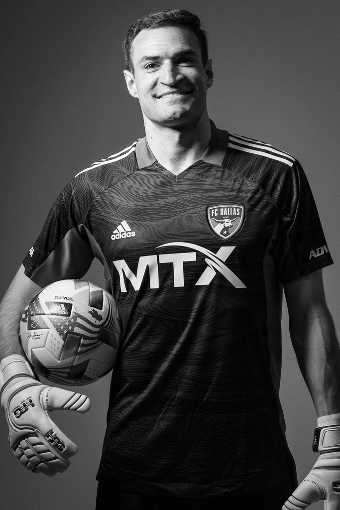 A monochrome photo of a soccer player wearing a dark FC Dallas jersey and gloves, holding a soccer ball under his arm and smiling, exuding the quiet influence of an experienced leader on the field.