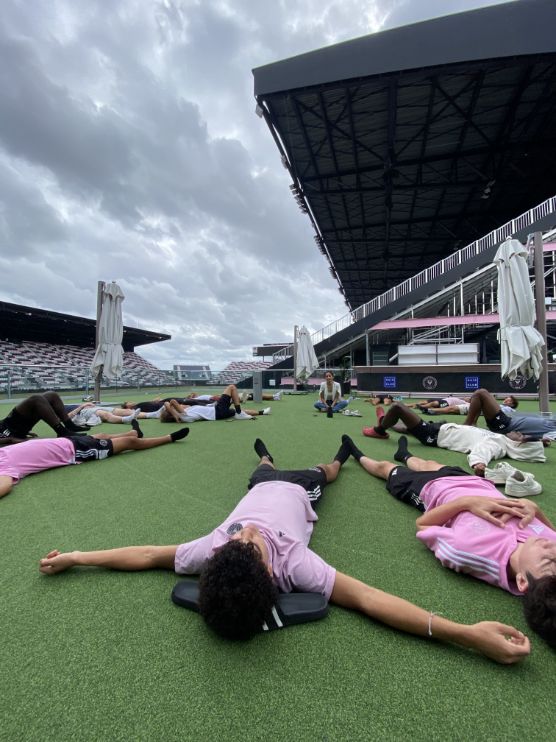 Soccer players from Inter Miami CF lie on artificial turf at a stadium, cooling down or practicing mindfulness under a cloudy sky. Stadium seating and structures loom in the background.