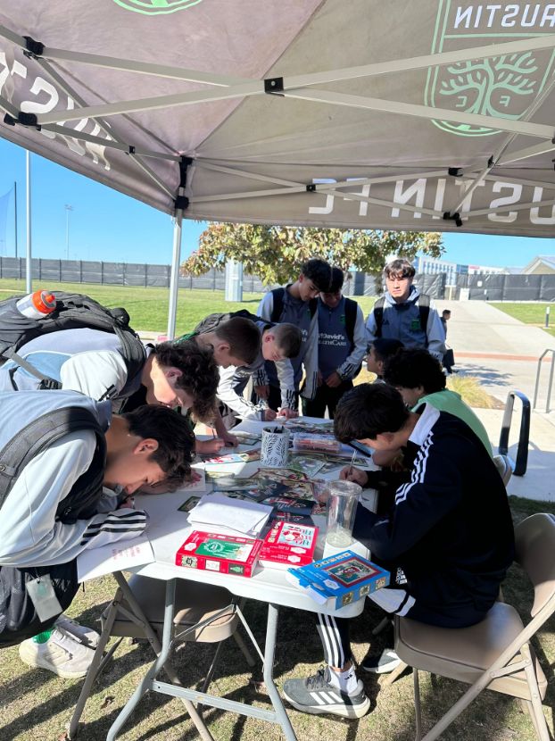 A group of people wearing athletic attire, including some Austin FC jerseys, gather under a canopy at a table filled with books and cards. They appear to be reading and writing. Some stand, while others sit at the table.