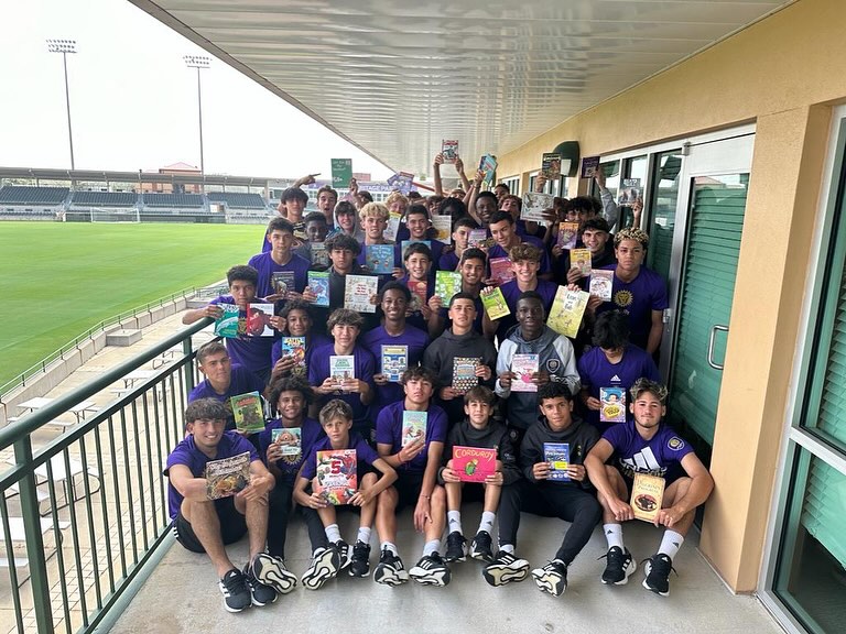 A large group of young people in sports uniforms stands on a stadium terrace, proudly holding up various children's books for display. They appear to be participating in a book collection or donation event in collaboration with Orlando City SC.