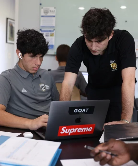 In a classroom, a teacher and a student with earbuds look intently at a laptop screen adorned with GOAT and Supreme stickers, possibly discussing financial aid opportunities.