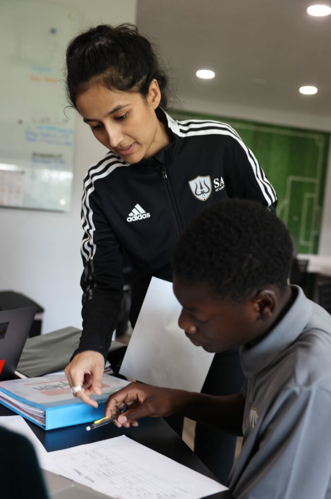 A woman in a black adidas jacket assists a seated student with his work at a desk on the SAI Seminole Campus.