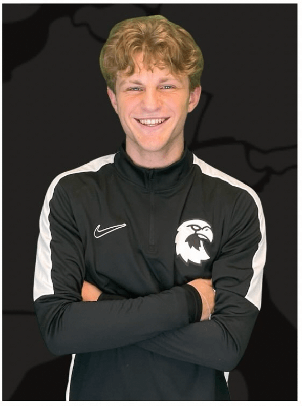 A person with curly hair is smiling and standing with arms crossed. They are wearing a black and white sports jacket with a bird logo and a Nike emblem, part of the Talentprojekt Campus collection. The background is a dark, indiscernible pattern.