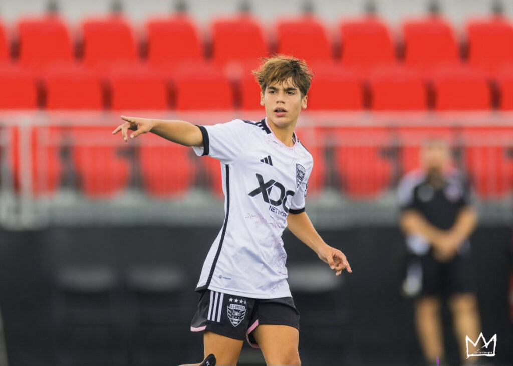 A soccer player in a white and black uniform points ahead on the field, with empty red seats in the background, reminiscent of a game day at the DC United Campus.