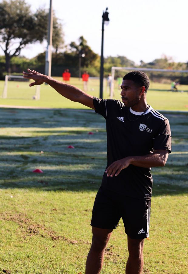 A man wearing a black soccer kit with a white logo gestures with his arm while standing on a grassy field. Soccer goals and other players are visible in the background, capturing the essence of the sport.