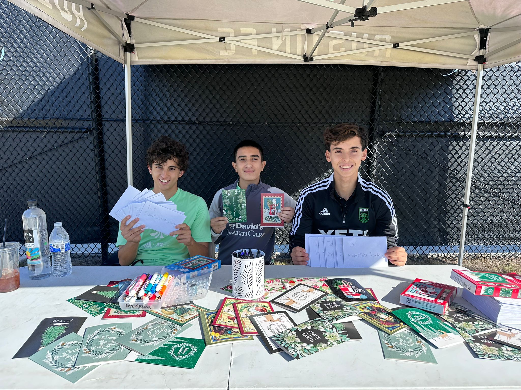 Three young men sit at a table under a canopy on the Austin FC Campus, displaying handmade cards. The table is covered with craft supplies, water bottles, and more cards. A black chain-link fence is visible in the background.