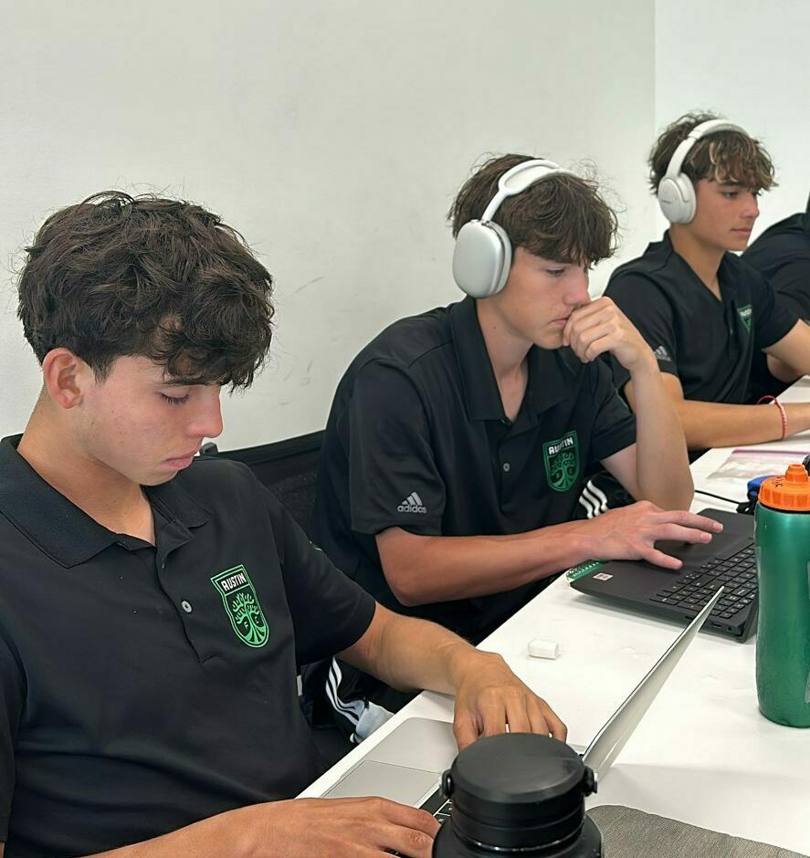 Three young men wearing headphones and matching black shirts are seated at a table on the Austin FC campus, focusing intently on their laptops. One has a green water bottle, and there is a minimalistic background.