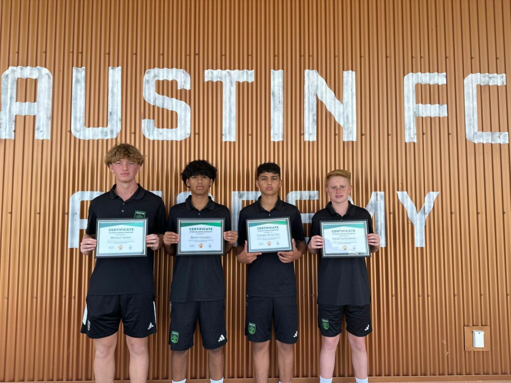 Four young athletes in black sportswear hold certificates in front of a wall with "Austin FC Academy" signage, celebrating their achievements at the Austin FC Campus.