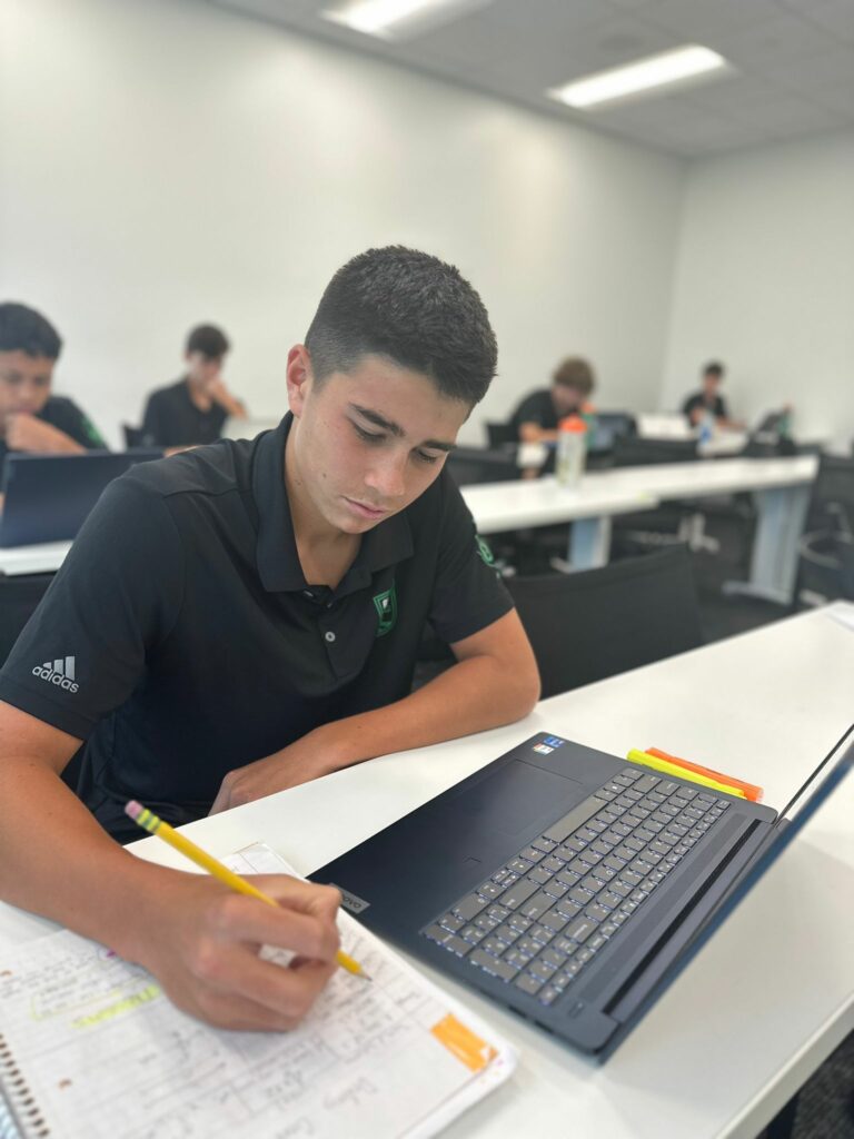 A person in a classroom writes notes on a notebook while using a laptop, displaying an Austin FC sticker. Other individuals are visible in the background, also focused on their work, embodying the studious atmosphere of the campus.