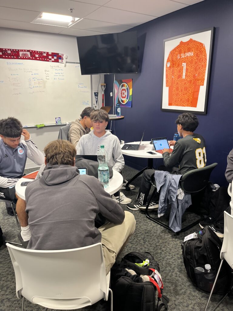 Several people are seated at tables in a room, using laptops and working. A sports jersey from the Chicago Fire Campus is framed on the wall, and there is a television and some decor on the walls.