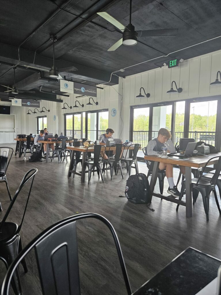 A spacious room on the DC United Campus features several individuals working on laptops at separate tables. Large windows allow natural light to enter, and ceiling fans are visible above. Backpacks are placed beside some chairs.