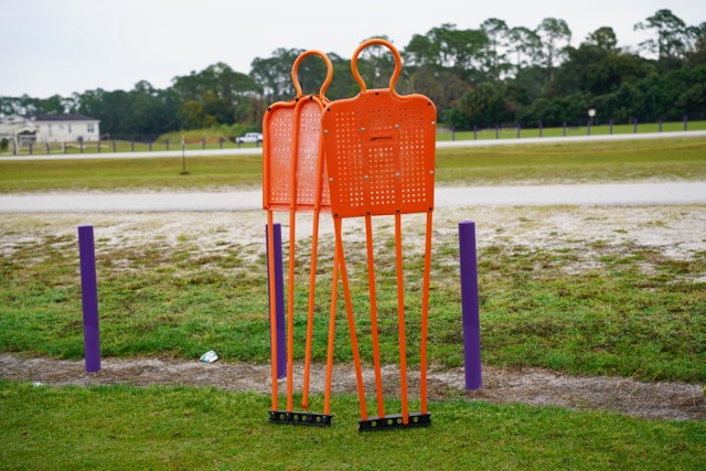 Three orange training mannequins stand on a grassy field with purple poles and a distant background of trees and cloudy sky.