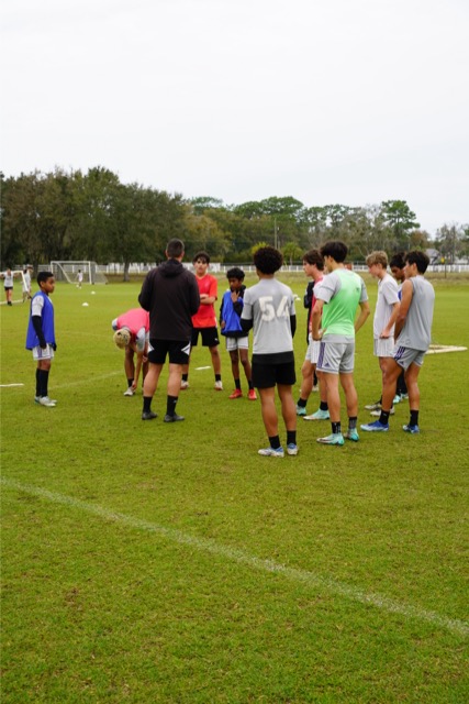 A group of young athletes, wearing sportswear and standing on a grassy field, gathers around two coaches who are demonstrating something. Soccer goals and more players can be seen in the background.
