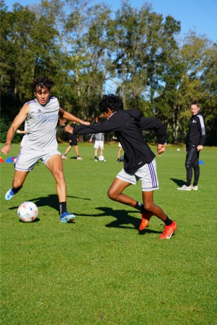 Soccer players in training on a grassy field, with one player dribbling the ball and another attempting to defend. A coach and other players are visible in the background among trees.