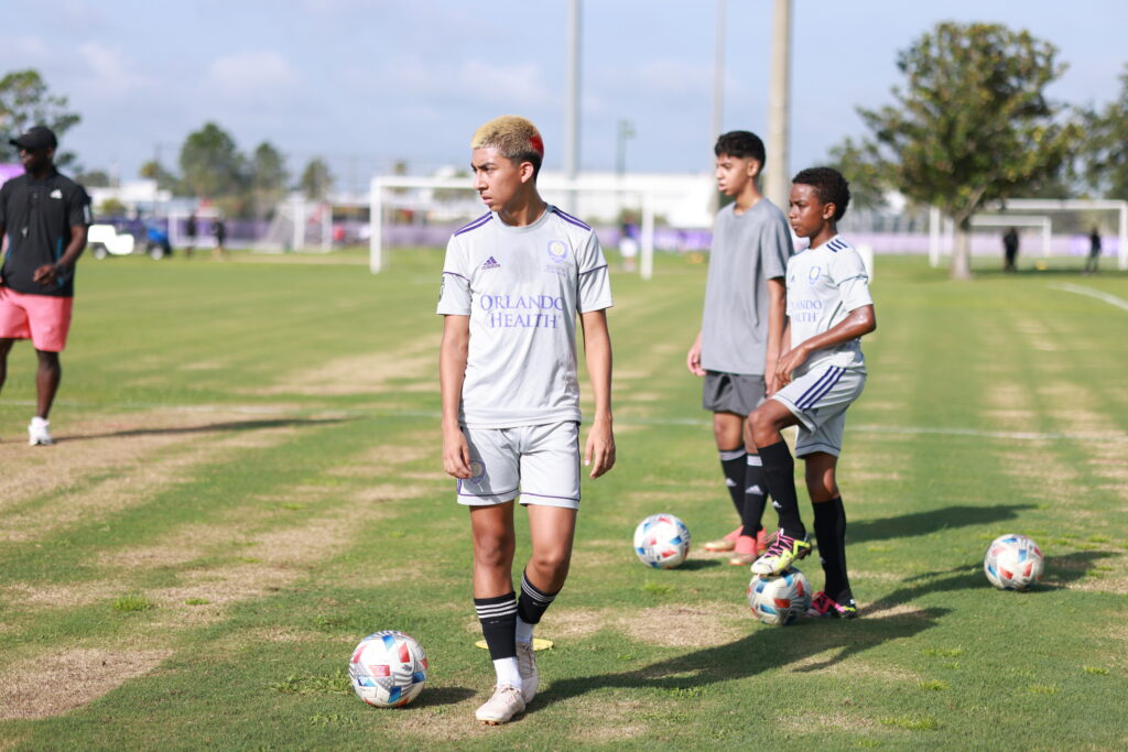 Young soccer players in gray uniforms stand on a grass field near soccer balls, preparing for practice at the Orlando City Campus. An adult in the background observes. Trees and other players can be seen in the distance.