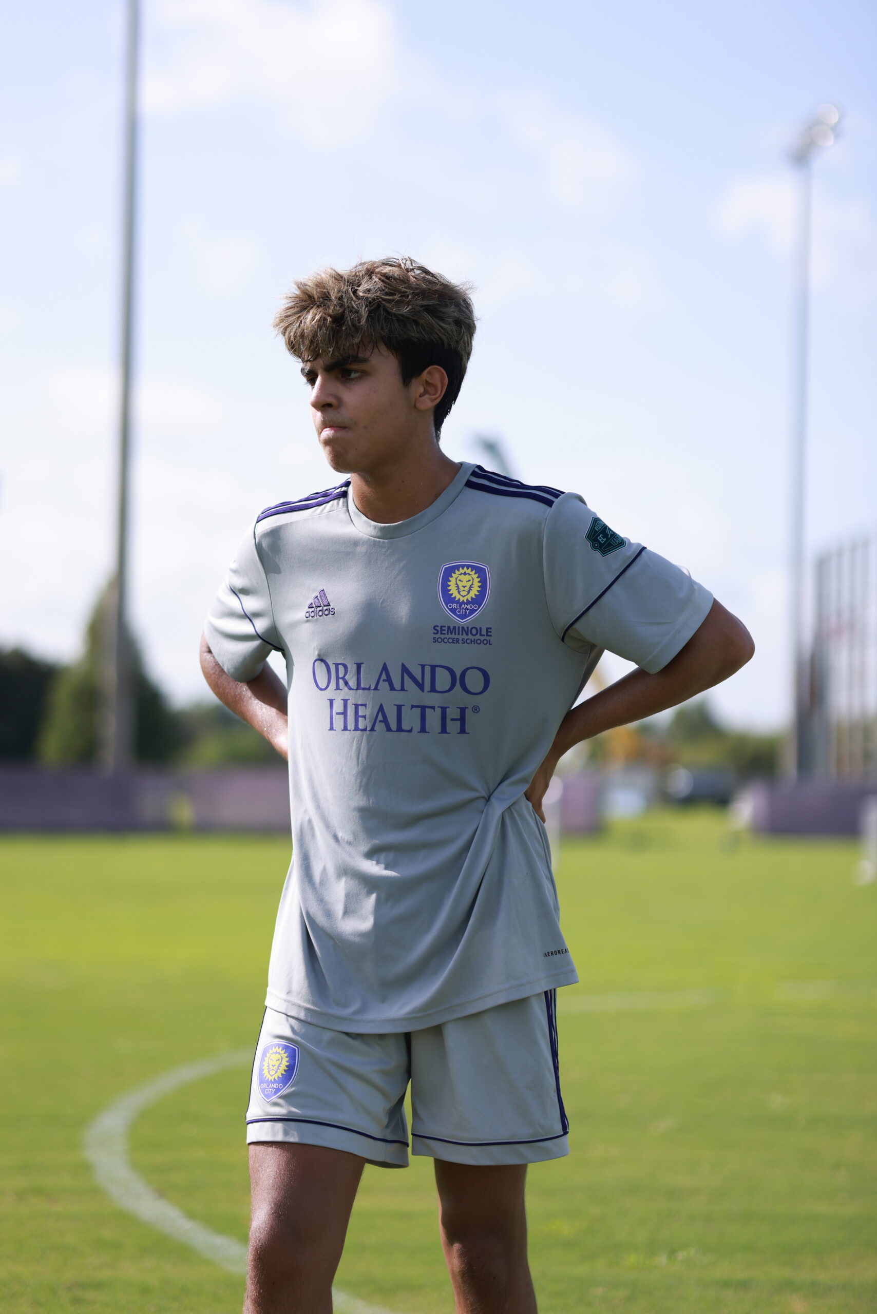 A soccer player in a light gray uniform with the logo "Orlando Health" stands on the Orlando City Campus' grassy field with hands on hips, looking to the side under a sunny sky.