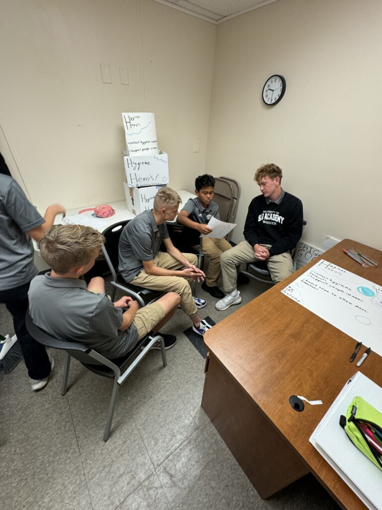 Four people are seated in a small room, engaged in a discussion. A whiteboard with notes rests on a table, and boxes are stacked in the corner. One person is holding a piece of paper, perhaps as they brainstorm ideas for the homepage redesign.