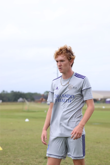 A person with curly hair wearing a gray "Orlando Health" sports uniform stands on a grassy field at SAI Academy.