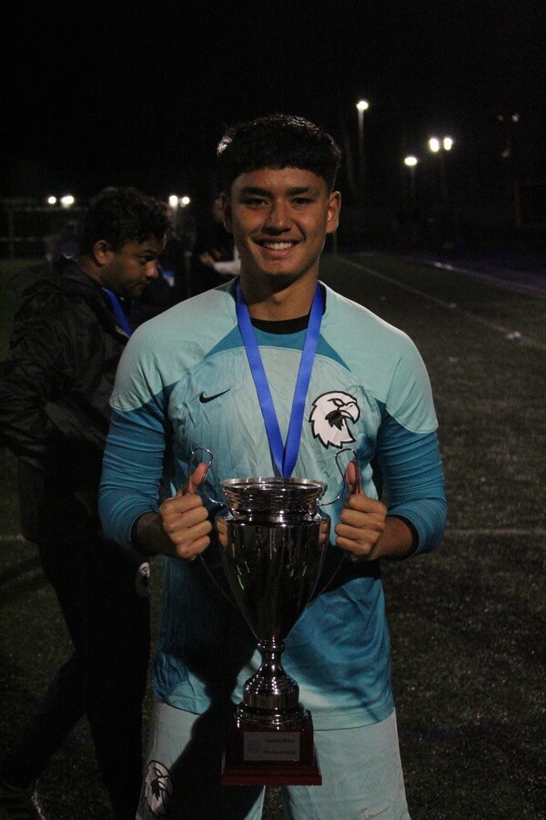 A soccer player in a blue uniform smiles while holding a large trophy at the Talentprojekt Campus. He has a medal around his neck, and another person is visible in the background.