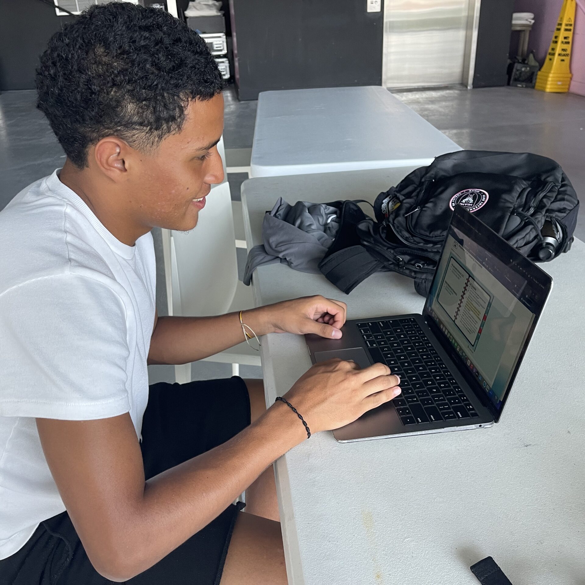 A person sits at a table on the Inter Miami Campus, using a laptop with a backpack and water bottle nearby.