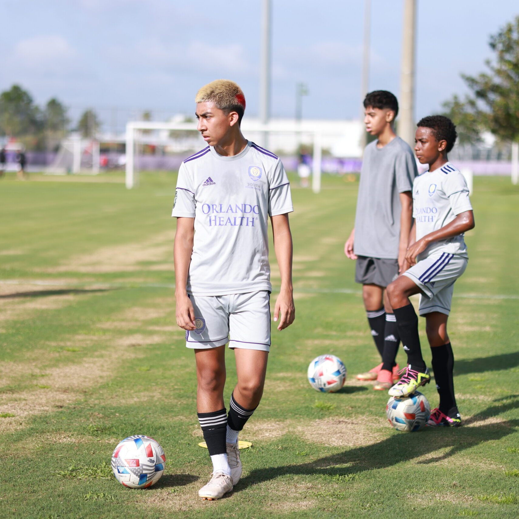 Young soccer players in gray uniforms stand on a grass field near soccer balls, preparing for practice at the Orlando City Campus. An adult in the background observes. Trees and other players can be seen in the distance.