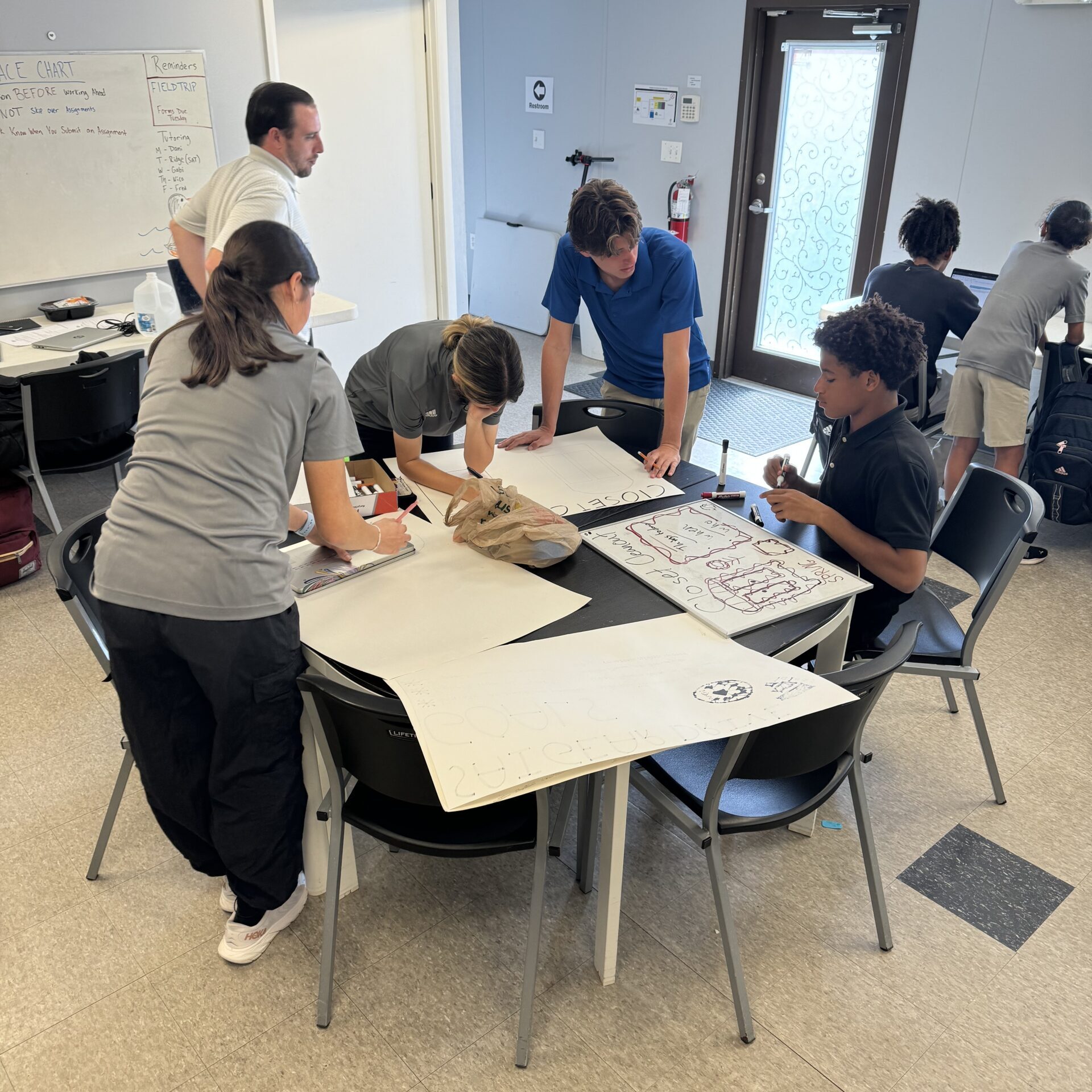 A group of people stands around a table covered with papers and markers, working on posters in a room with white walls and a door in the background, brainstorming ideas for their website's homepage.
